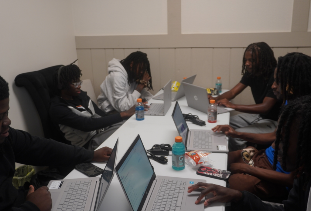 Group of young students sitting around a long white table working together on silver laptops in a classroom.
